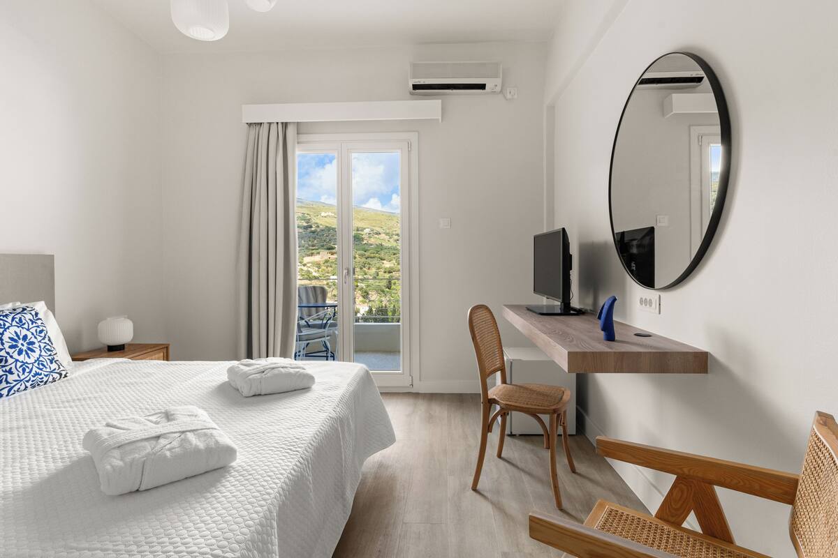 A hotel room with a bed, a desk, a chair, a TV, and a balcony with a view of the countryside.