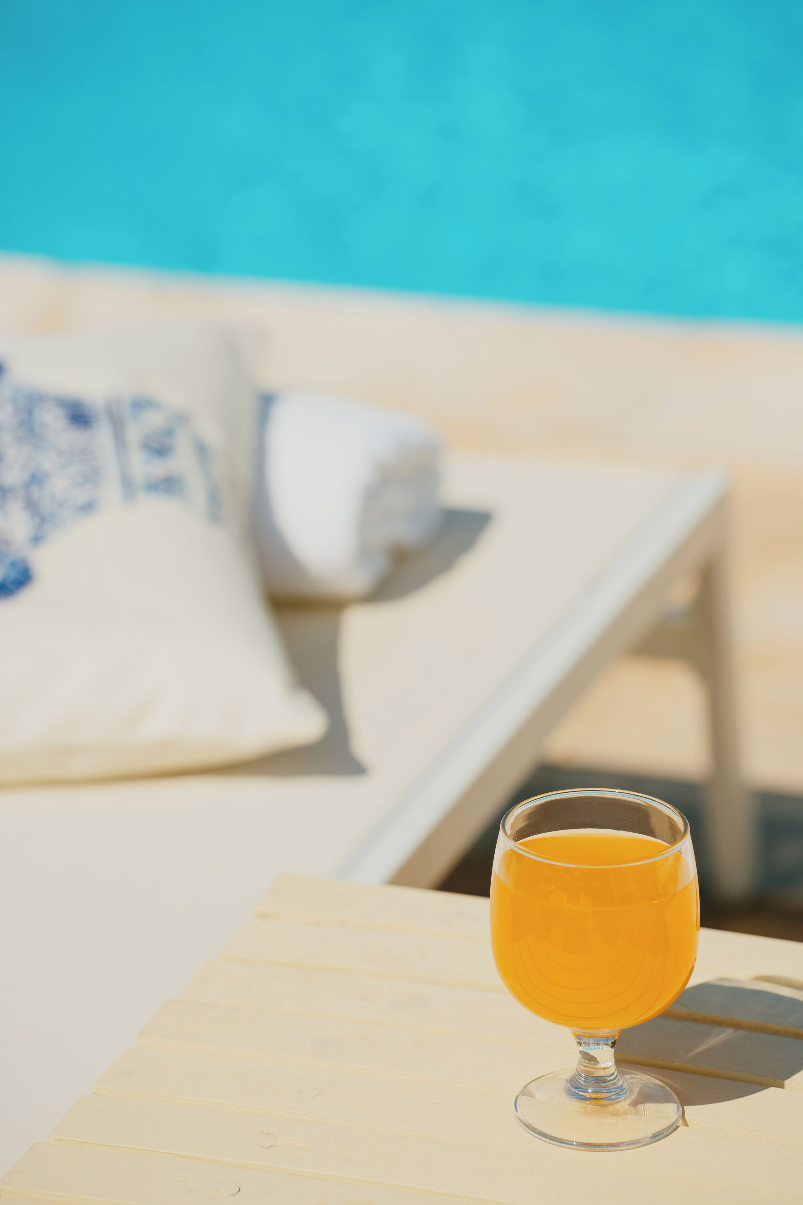 Un verre de jus d’orange sur une table en bois, à côté d’un fauteuil de relaxation placé au bord de la piscine, avec un coussin blanc.
