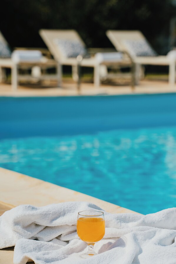 Un vaso de jugo de naranja sobre una toalla al lado de una piscina, con sillones de descanso al fondo.