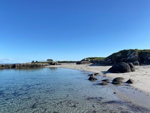 A beach with clear shallow waters, large rocks, and a grassy hill in the background.