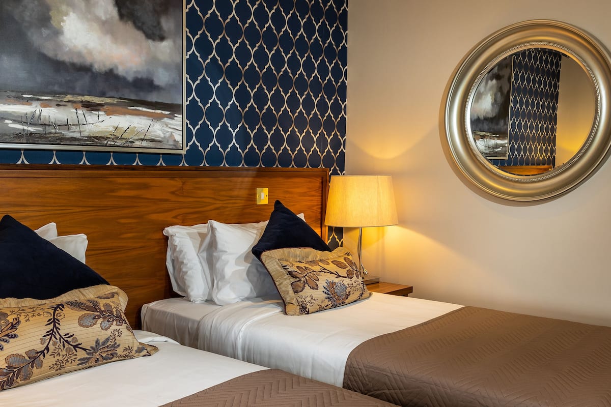 Une chambre d’hôtel avec deux lits, une tête de lit en bois, un grand tableau, un miroir rond et un mur décoré d’un motif.
