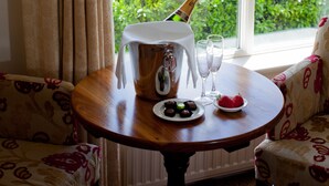 Een tafel met een champagnefles, twee glazen, een bord met aardbeien en een schaal chocolade bij een raam met uitzicht op groen en een waterpartij.