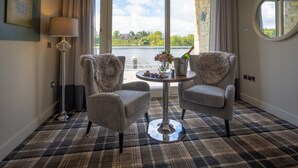 Een kamer met twee fauteuils, een tafel met een bloemenvaas, een fles champagne en uitzicht op een wateroppervlak door grote ramen.