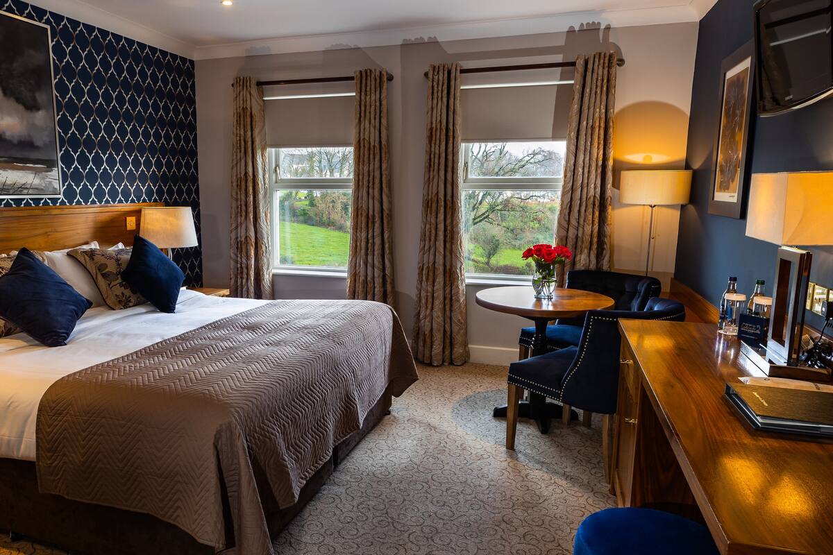 Une chambre d’hôtel avec un grand lit, un bureau avec une chaise, une fenêtre avec des rideaux et un tableau au mur.