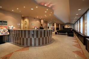 A hotel reception area with a curved counter, a man standing behind it, and a seating area with a view of the outside. - Astrid Centre Hotel (Brussels)