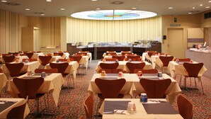 Een vergaderzaal met meerdere tafels en stoelen, een buffetgedeelte en een grote ronde plafondlamp.