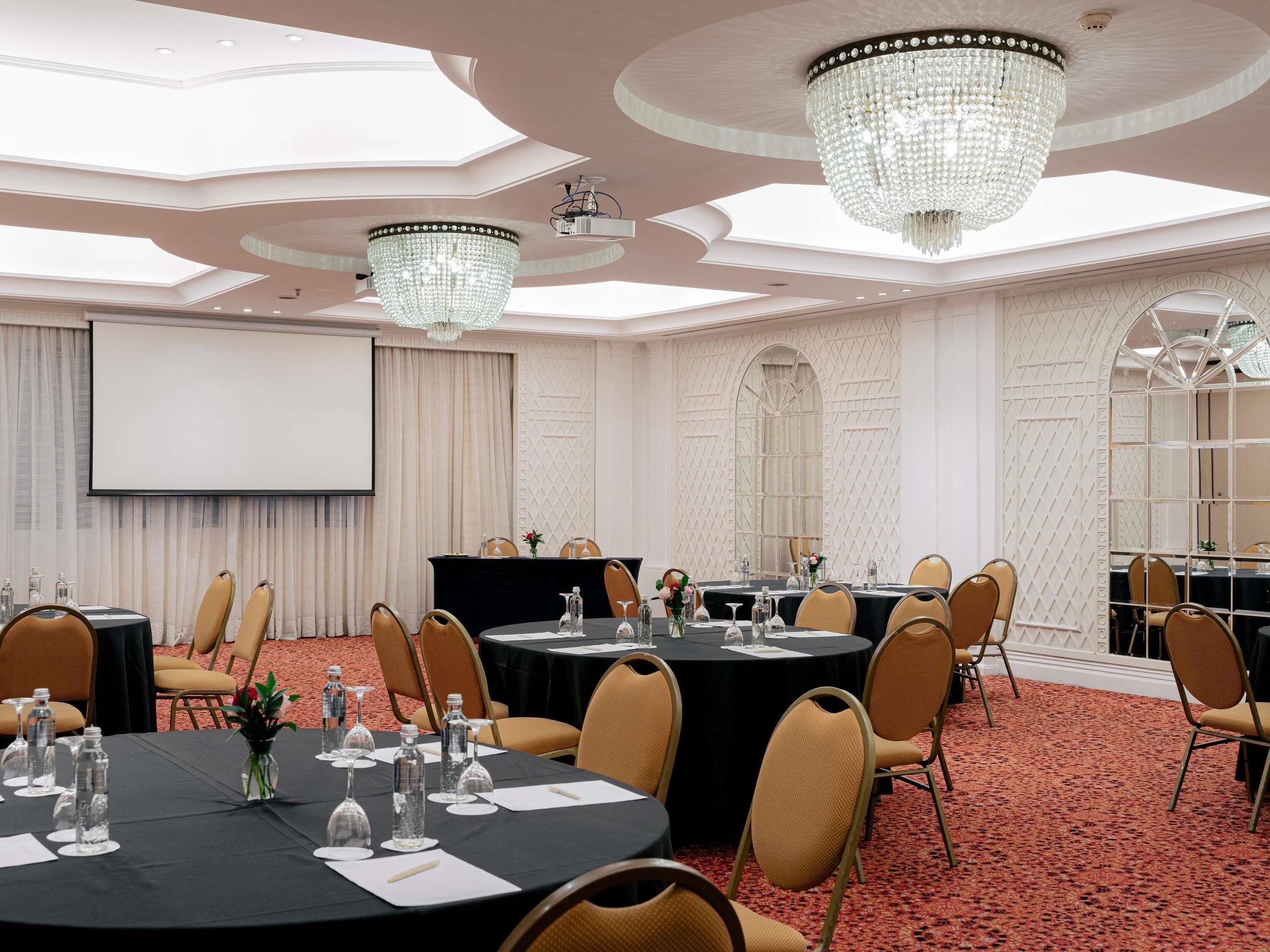 A conference room with round tables, chairs, a projector screen, and chandeliers.