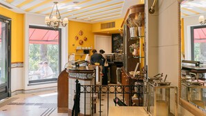 Interior de una cafetería con una decoración amarilla y blanca, un tablero de menú visible y una barra donde el personal está atendiendo.
