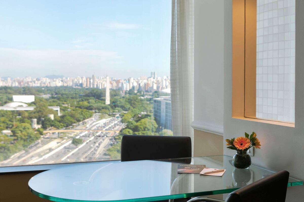 Habitación de hotel con una mesa de vidrio, un florero con flores y vistas a un paisaje urbano.