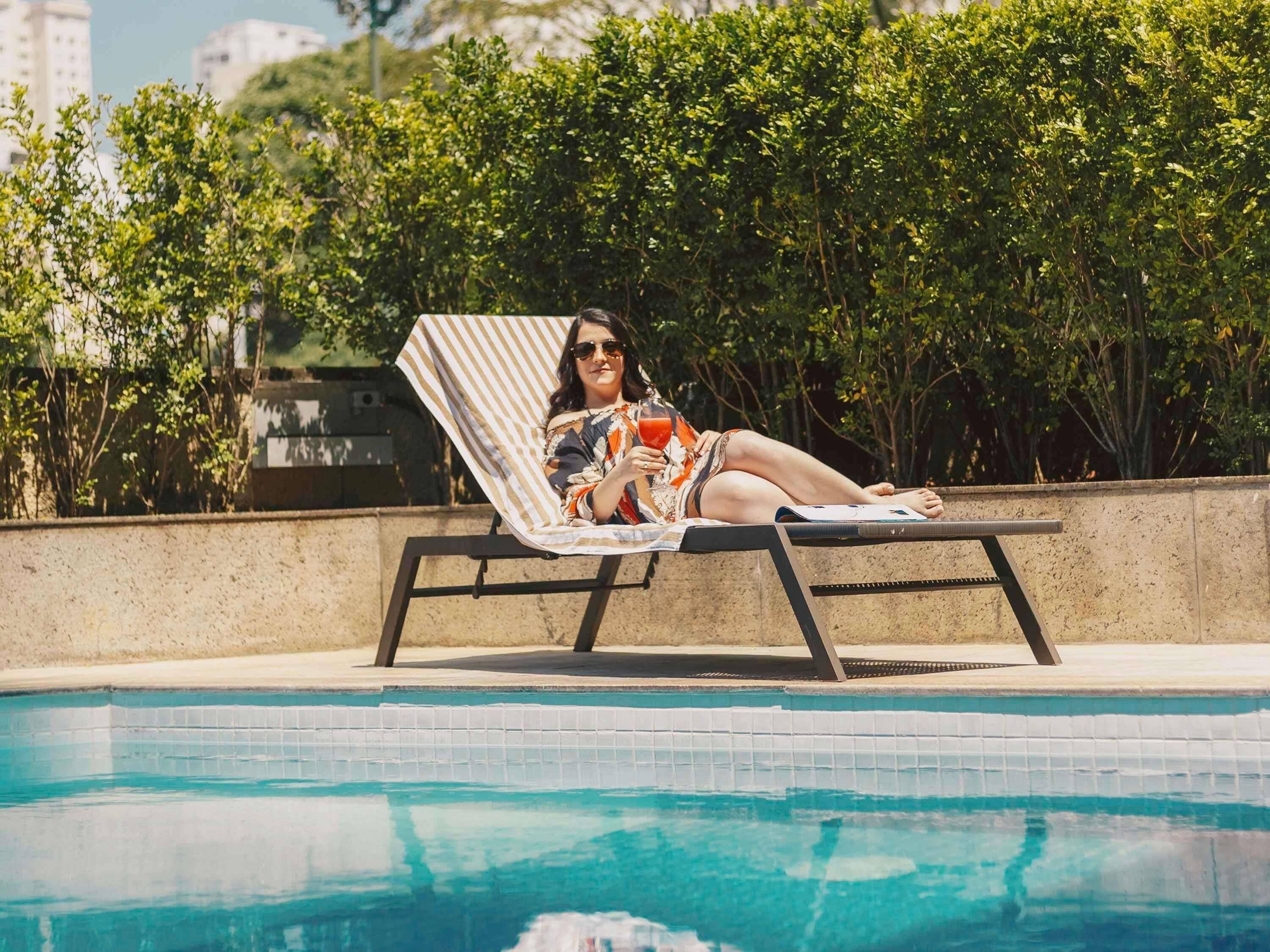 Una persona descansando en una tumbona junto a una piscina con borde de azulejos, rodeada de vegetación y edificios al fondo.