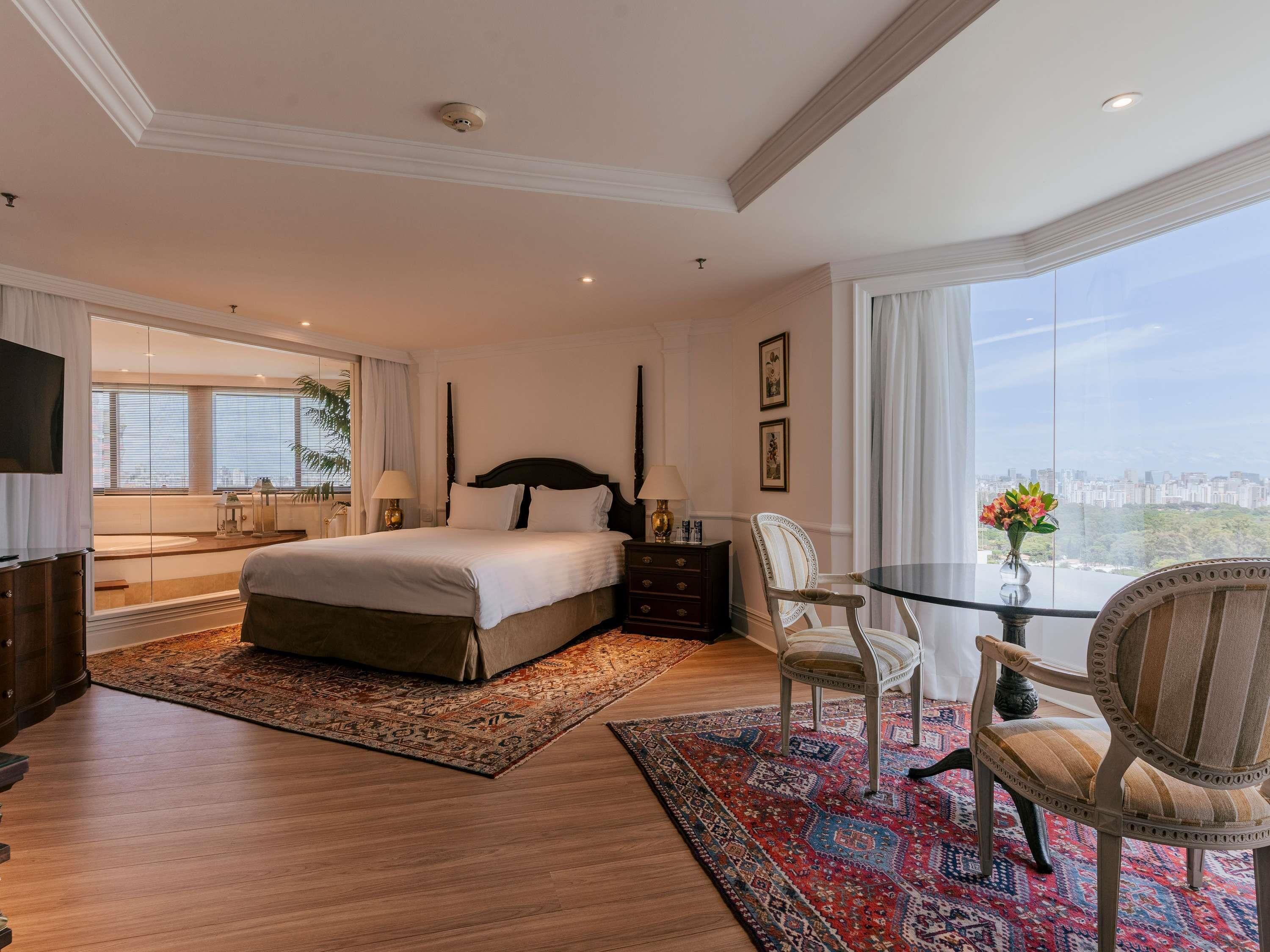 A spacious bedroom with a large bed, a glass table, two chairs, a nightstand, and a city view.