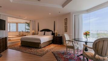A spacious bedroom with a large bed, a glass table, two chairs, a nightstand, and a city view.