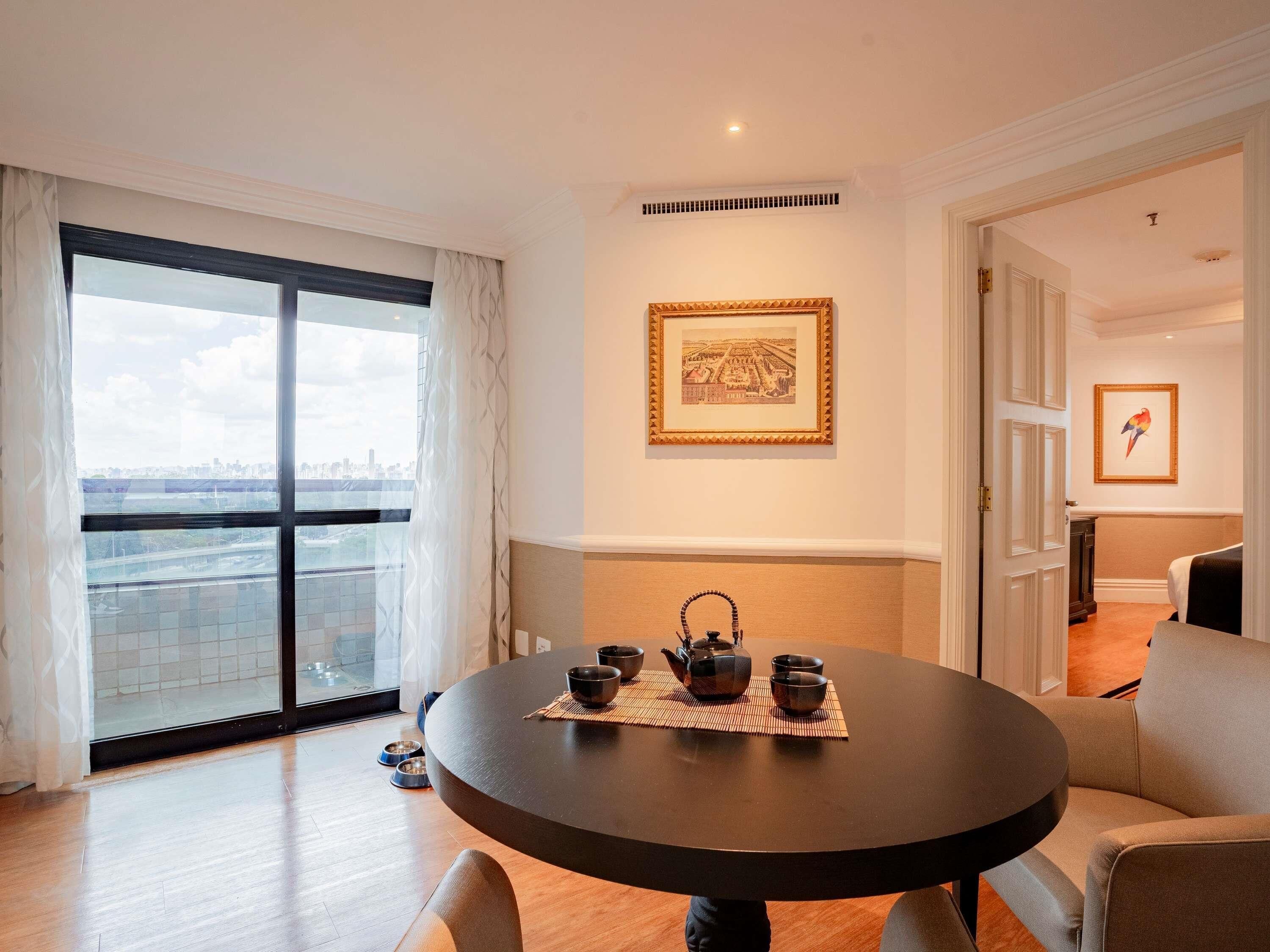 A modern living room with a round table set for tea, a large window with a city view, and a framed picture on the wall.