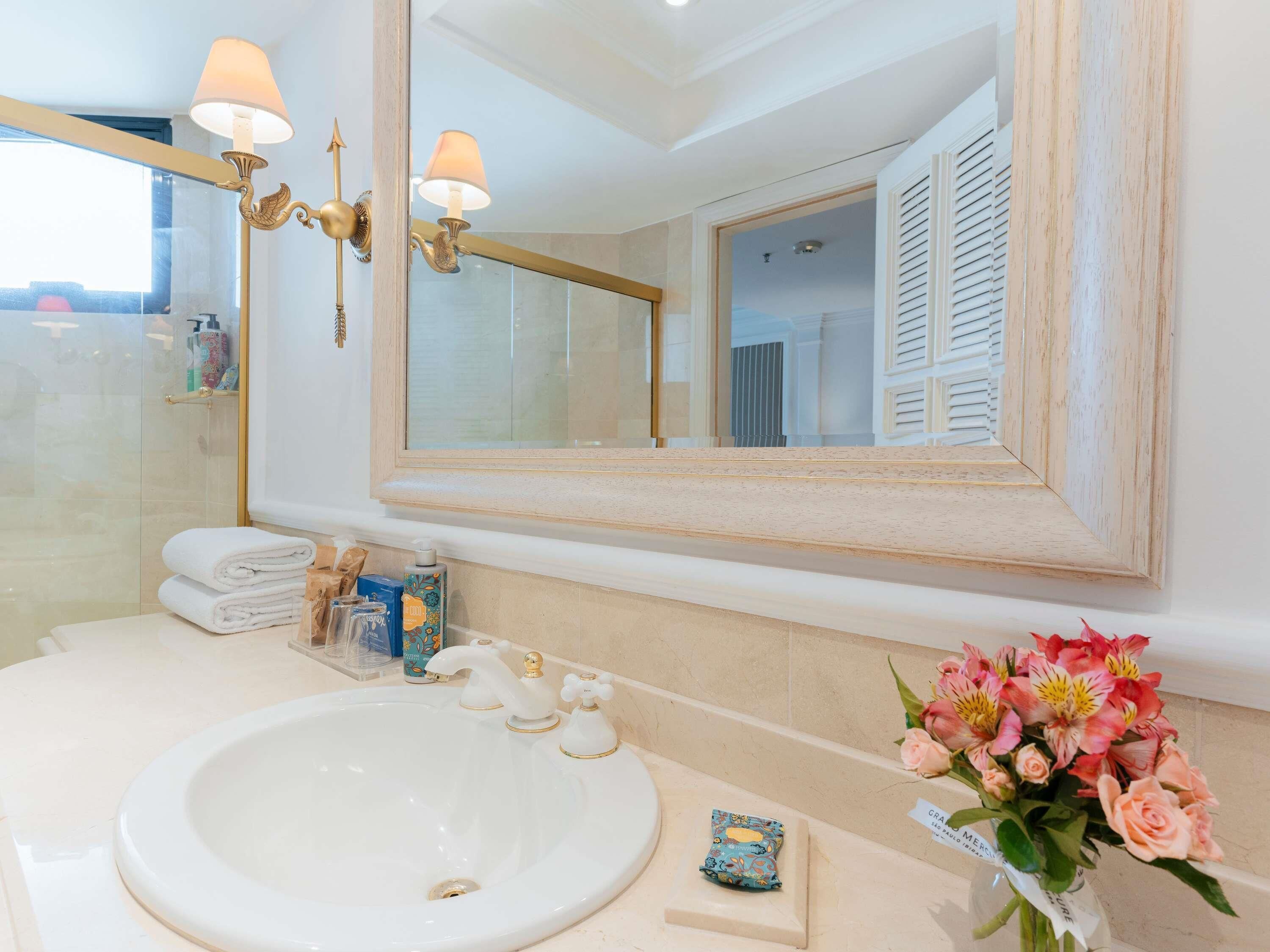 Un baño con un lavamanos blanco, dos lámparas de pared, un espejo grande y un florero con flores.