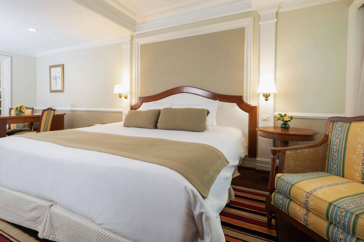 Habitación de hotel con una cama grande, dos mesitas de noche, una silla y vista al pasillo.