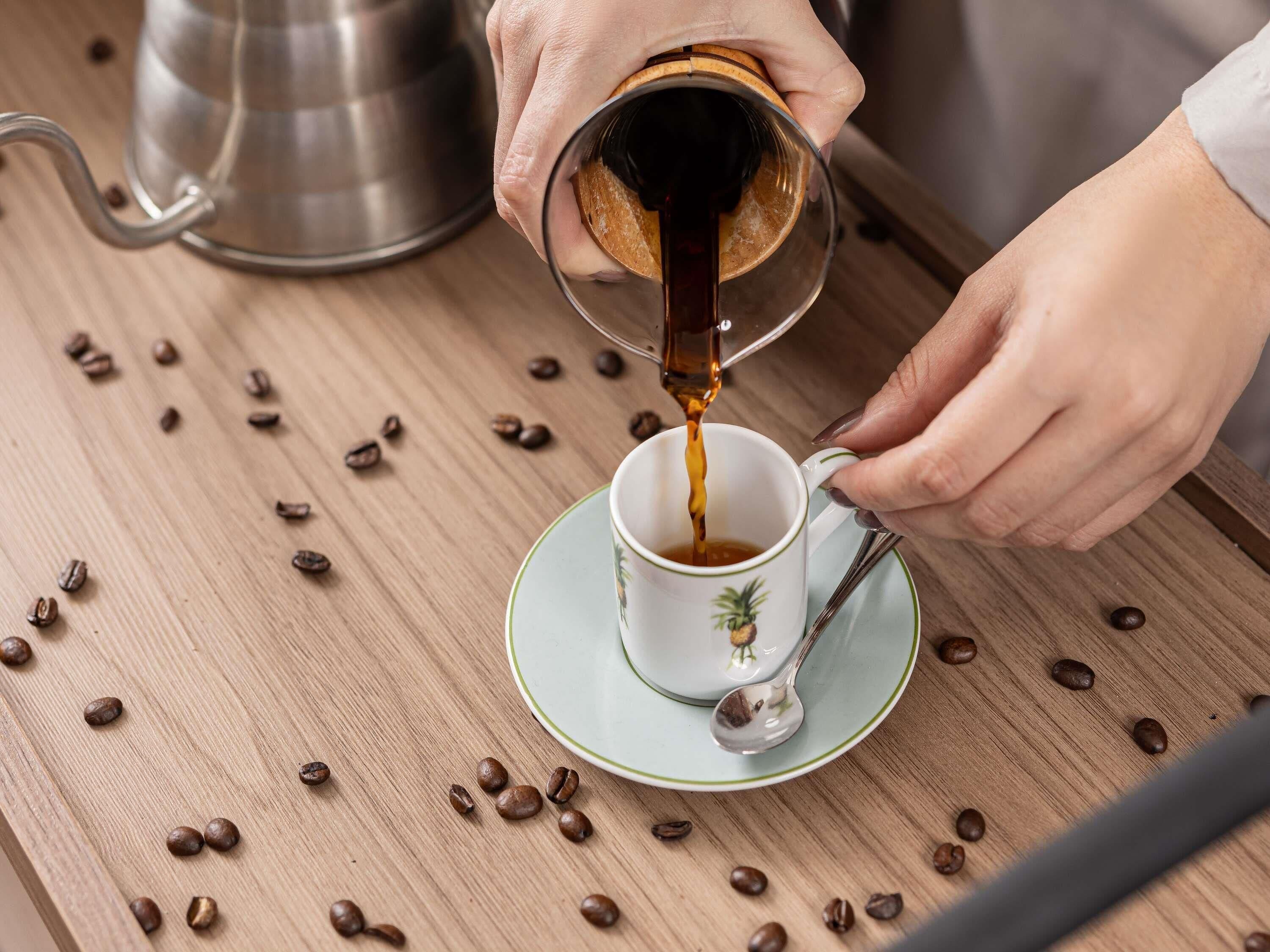 A person pouring coffee into a cup on a wooden table surrounded by coffee beans.