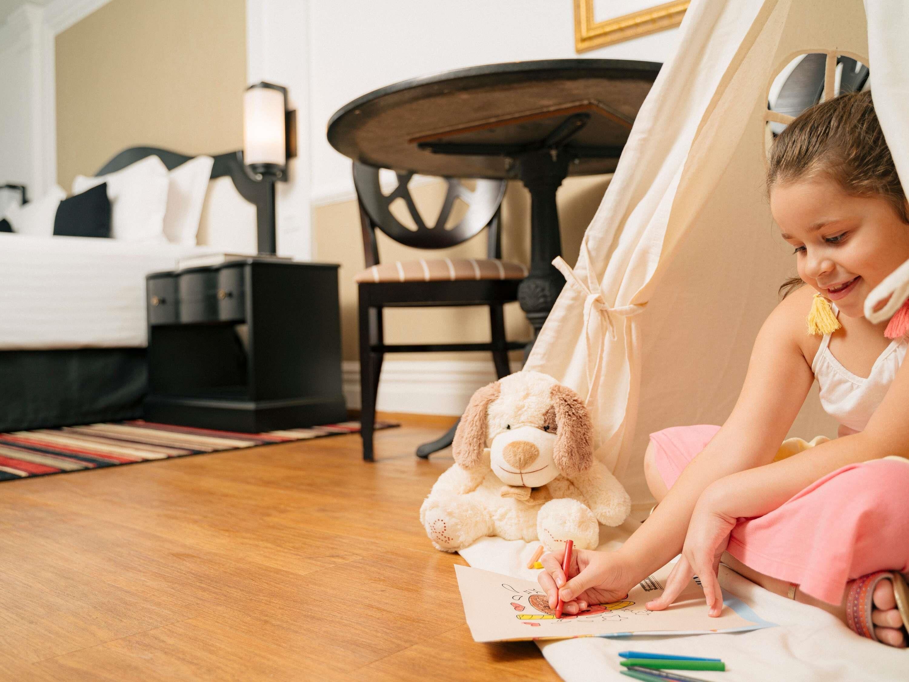 A child drawing at a wooden table with a tent in a room with a bed and a lamp.