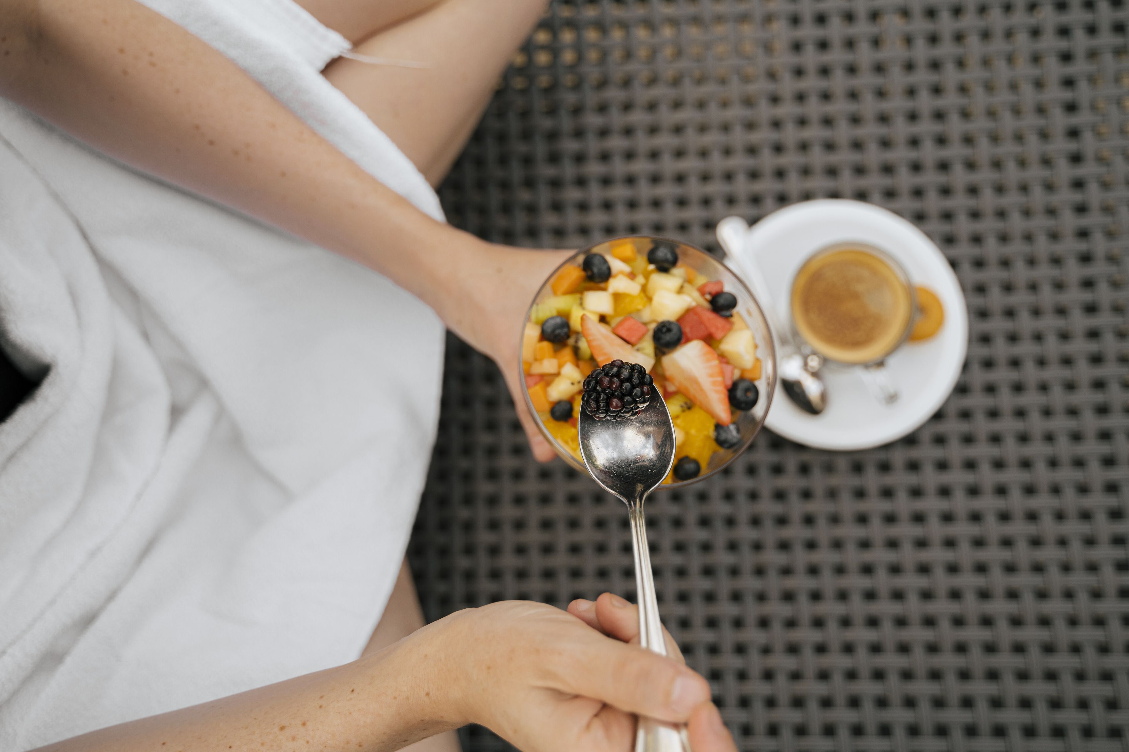 A person holding a spoon with a fruit salad and a cup of coffee on a table.