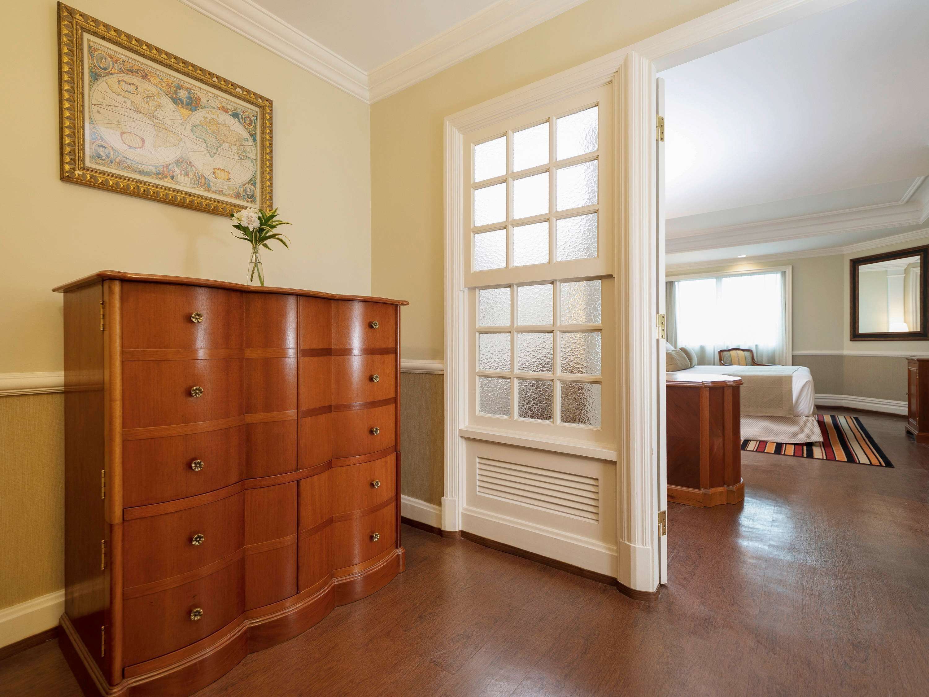 A room with a wooden dresser, a large window with frosted glass, and a bed with a striped rug.