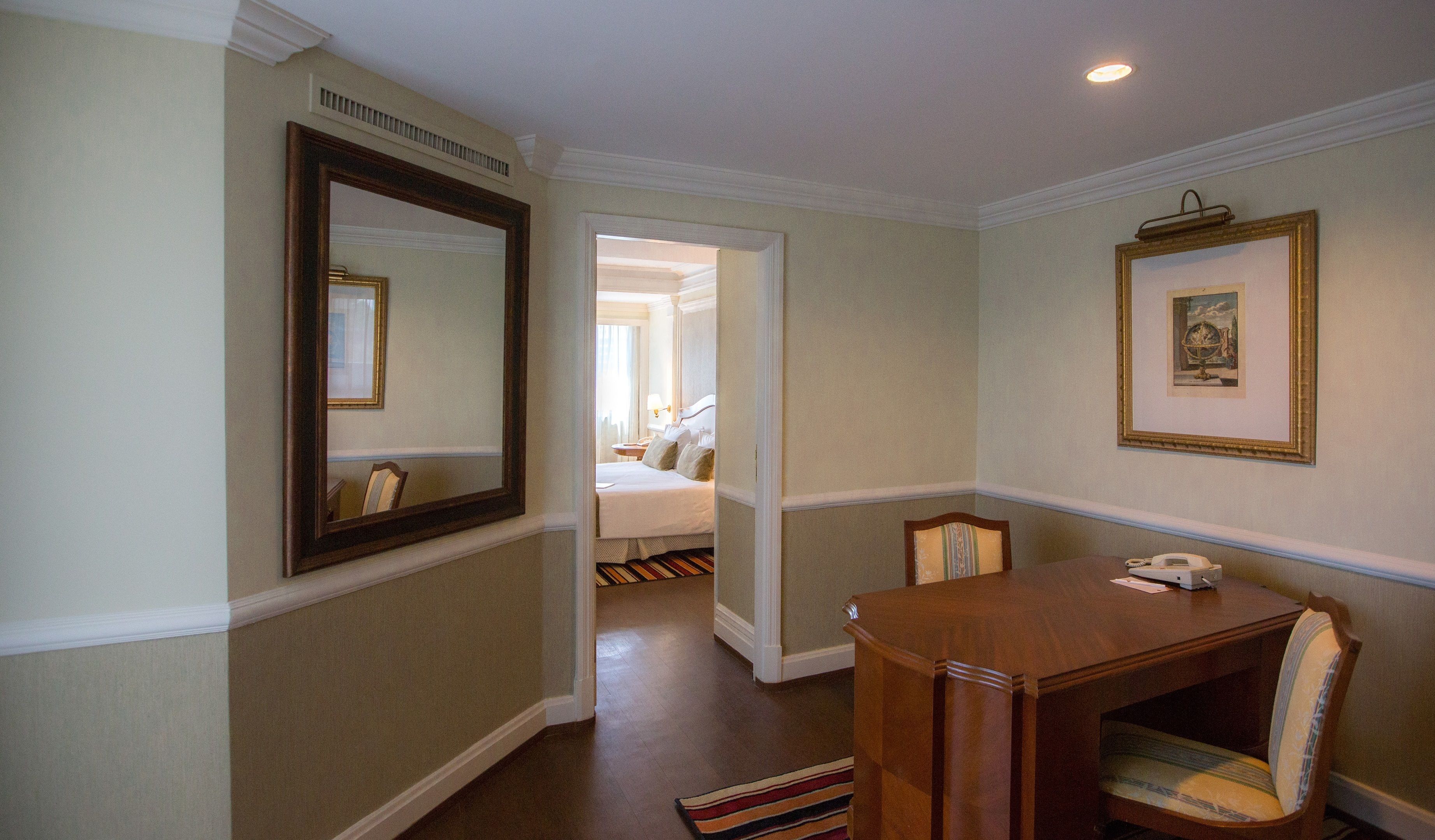 A hotel room with a large mirror, a dining table with chairs, a small desk with a telephone, and a framed picture on the wall.
