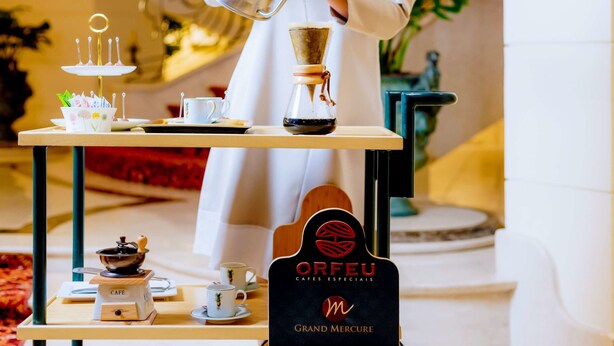 Una persona sirve café en una taza en una cafetería, con un molinillo, una prensa francesa y una exhibición de tazas y platitos.
