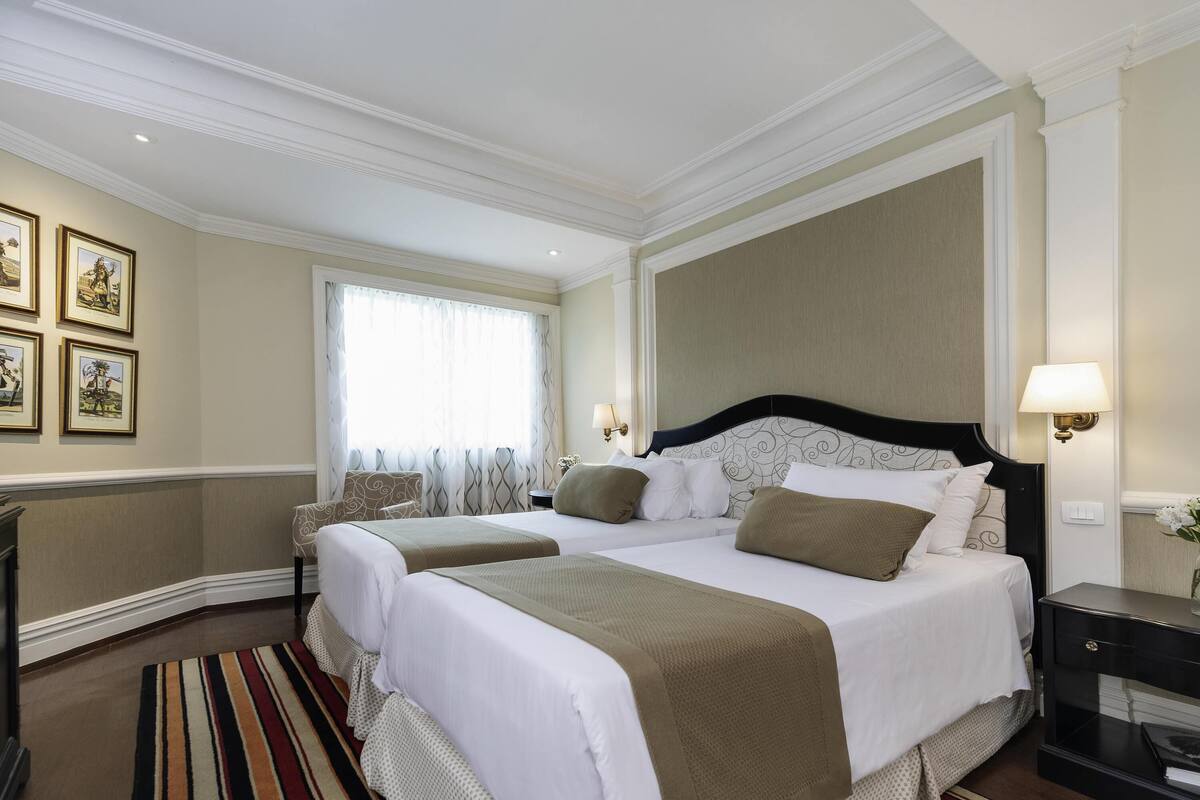 Habitación de hotel con dos camas, un ventanal con cortinas, una mesita de noche con lámpara y una alfombra estampada.