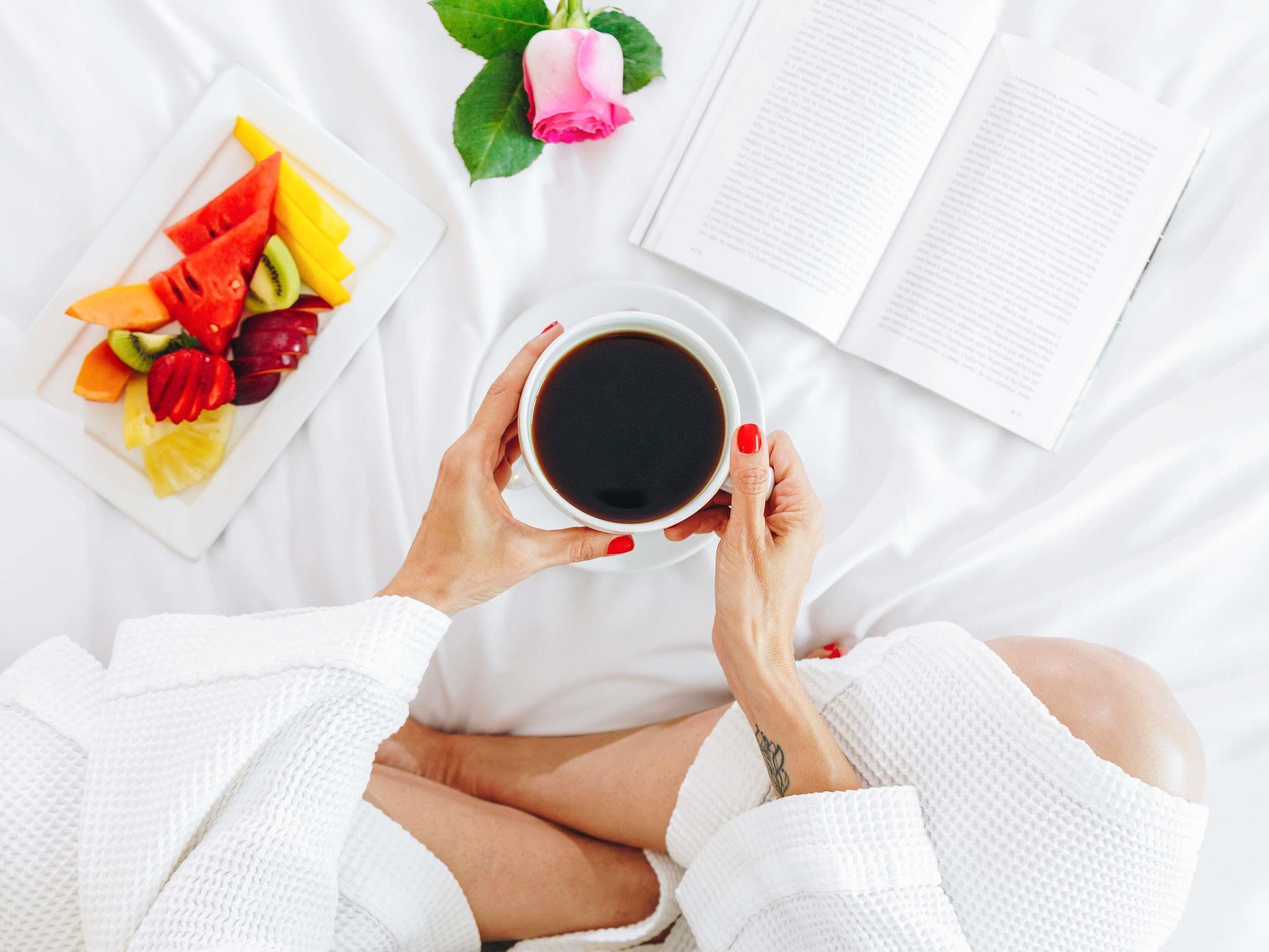 Une personne tenant une tasse de café, une assiette de fruits et un livre, le tout posé sur un lit blanc.