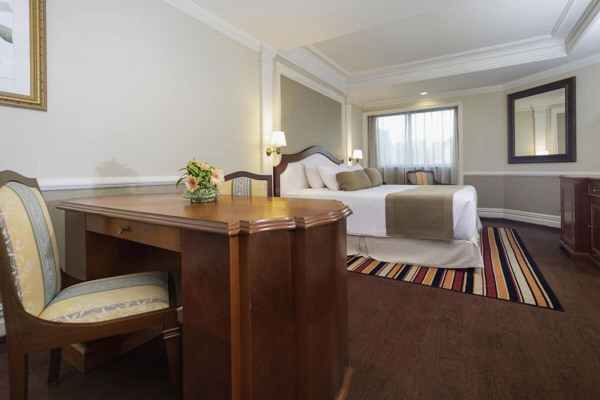 Habitación de hotel con cama, escritorio, silla, lámpara, espejo y ventana con cortinas.
