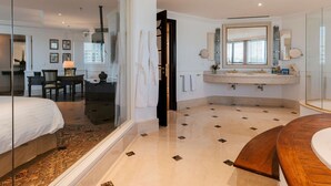 Una habitación de hotel moderna con una cama grande, una zona de estar con una mesa y sillas, un baño con tocador y espejo, y cabina de ducha.