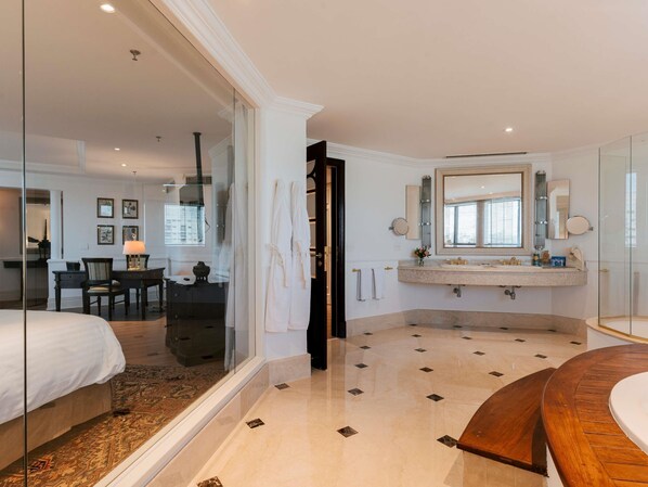 Una habitación de hotel moderna con una cama grande, una zona de estar con una mesa y sillas, un baño con tocador y espejo, y cabina de ducha.