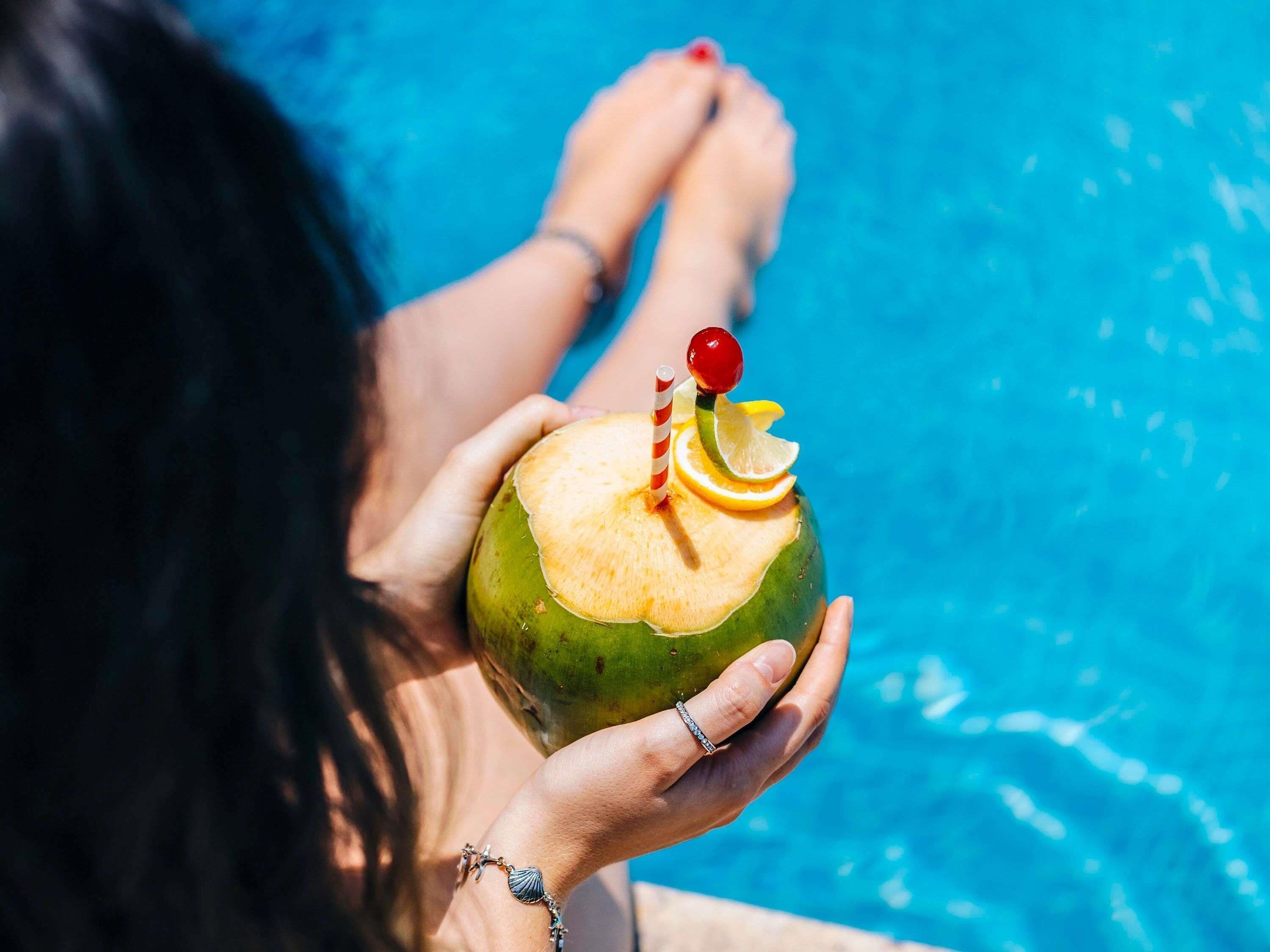 Une personne tenant une noix de coco avec une paille et une cerise sur le dessus, près d’une piscine.