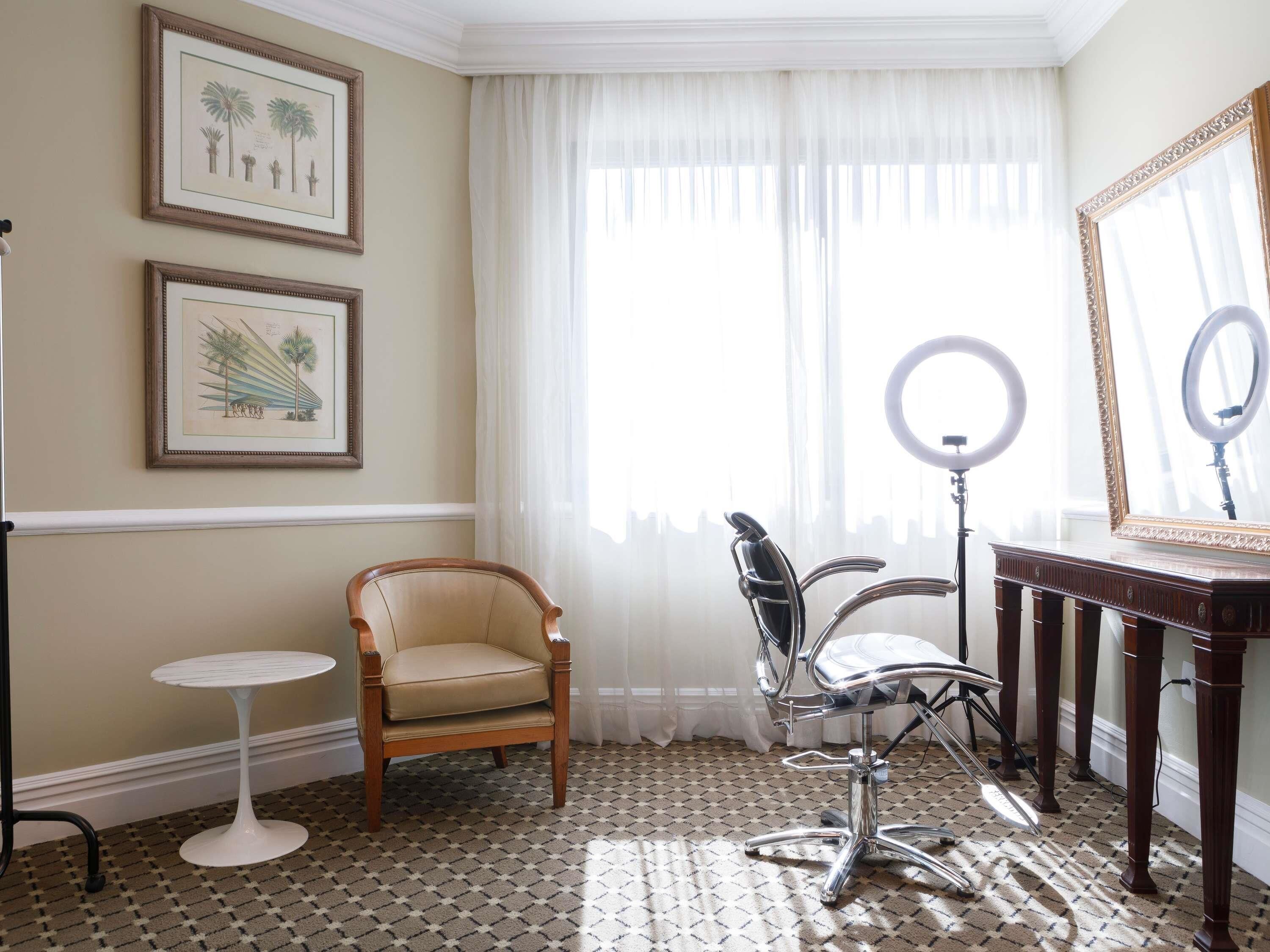 A room with a patterned carpet, a wooden chair, a modern chair, a small round table, and a mirror with a ring light.