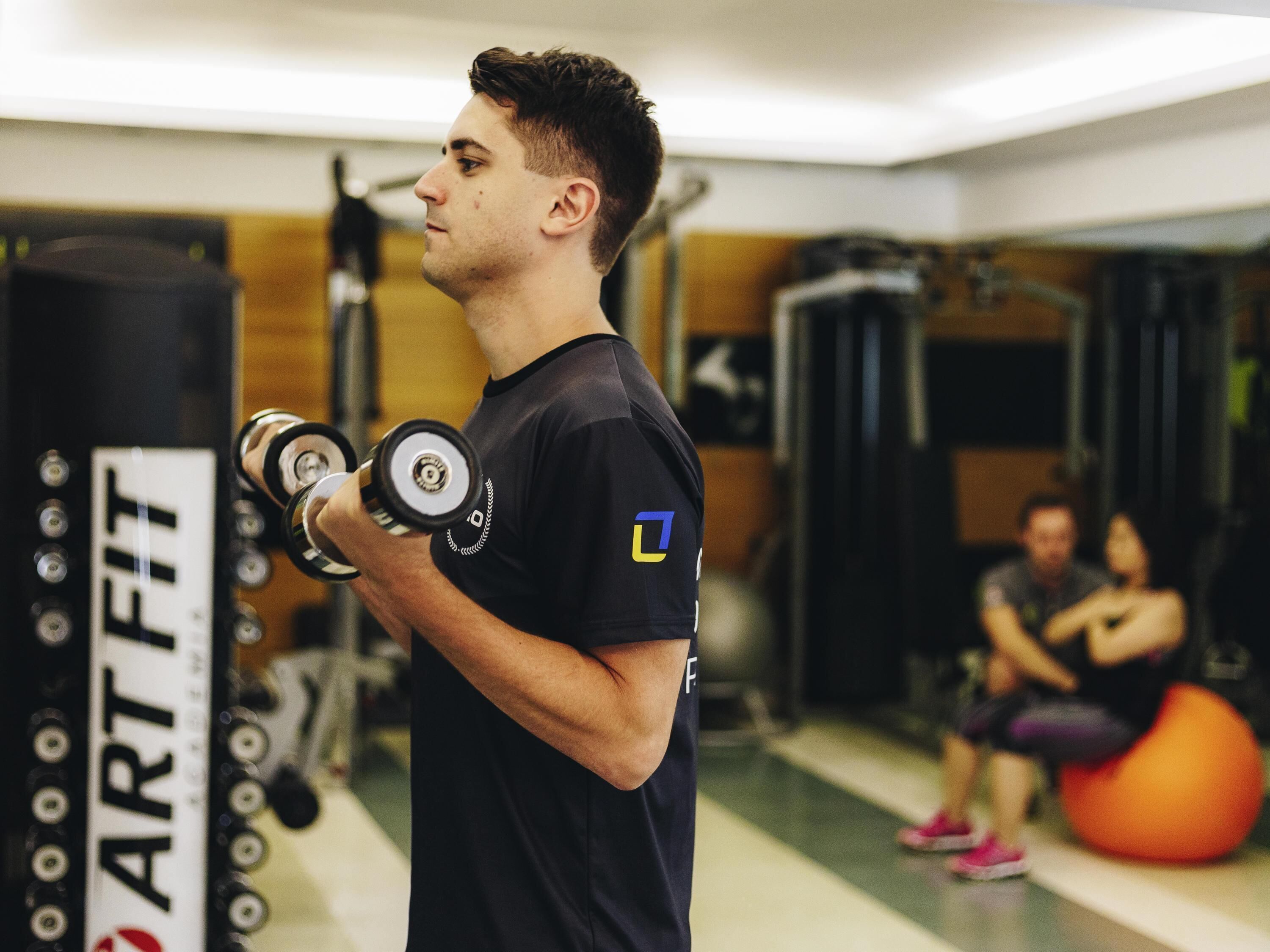 A gym with exercise equipment, a person lifting weights, and another person sitting on an exercise ball in the background.