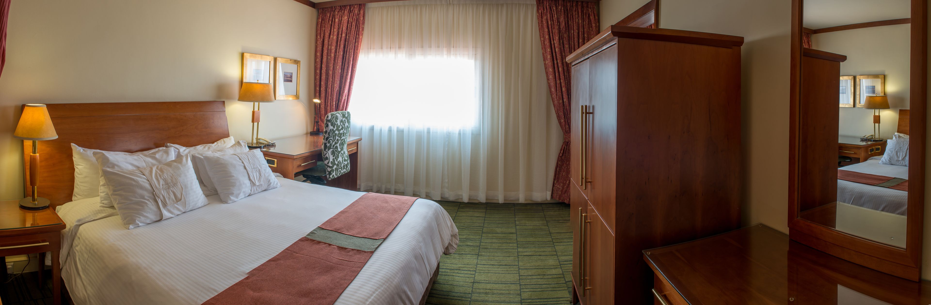 A hotel room with a large bed, a wooden wardrobe, a desk with a lamp, and a mirror.