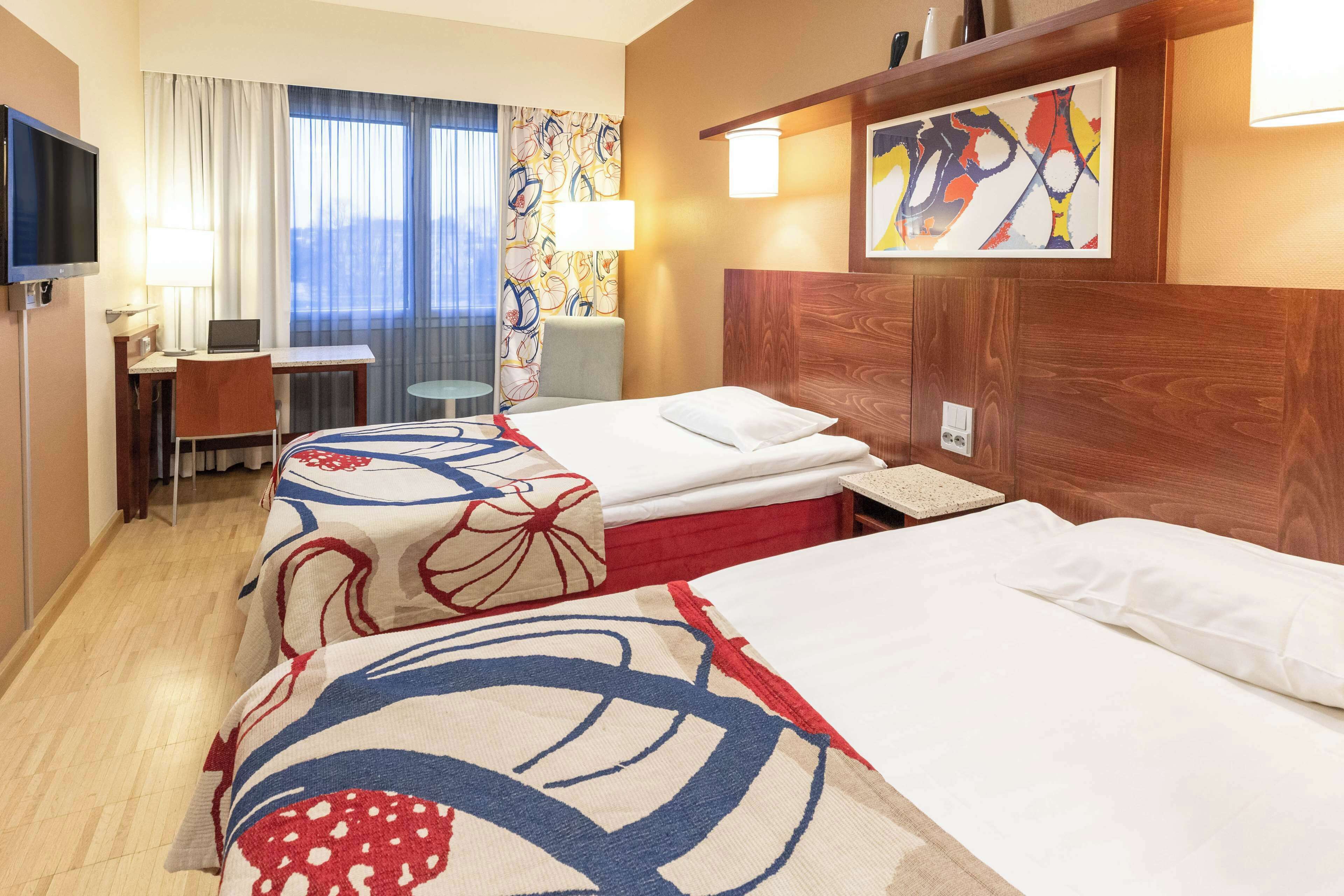 Hotellihuone, jossa on kaksi sänkyä, työpöytä kannettavalla tietokoneella, televisio ja ikkuna, jossa on verhot.