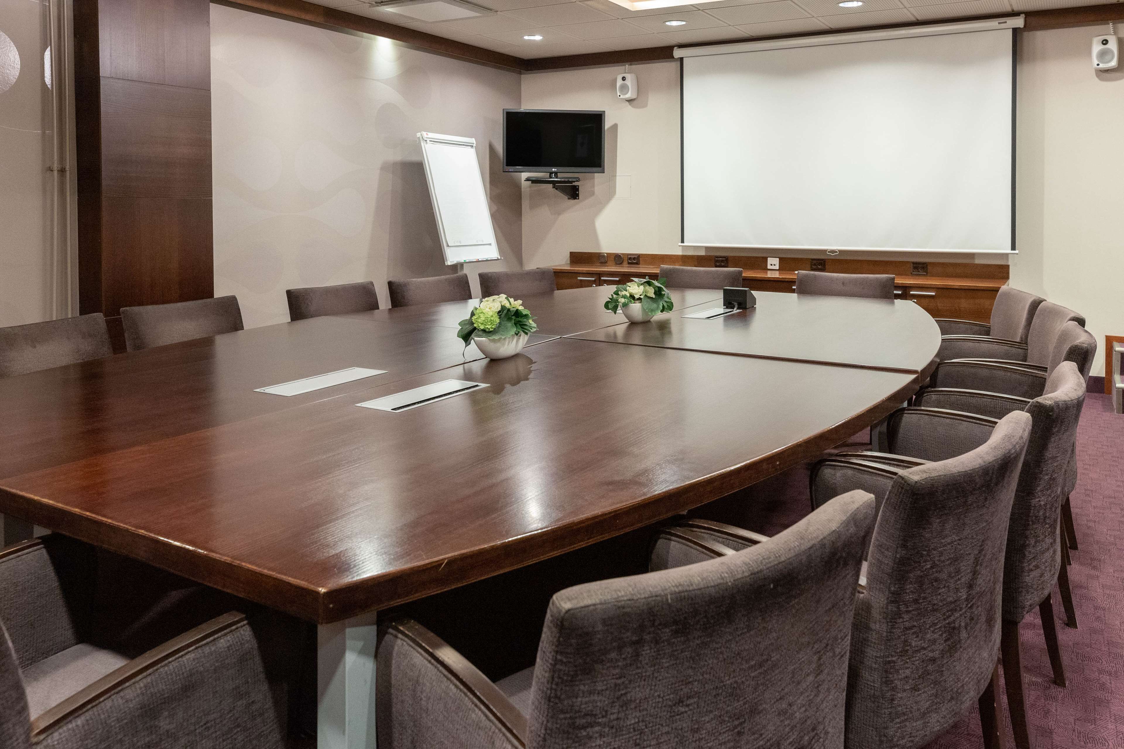 A conference room with a large wooden table, grey upholstered chairs, a whiteboard, a projector screen, and a television on the wall.