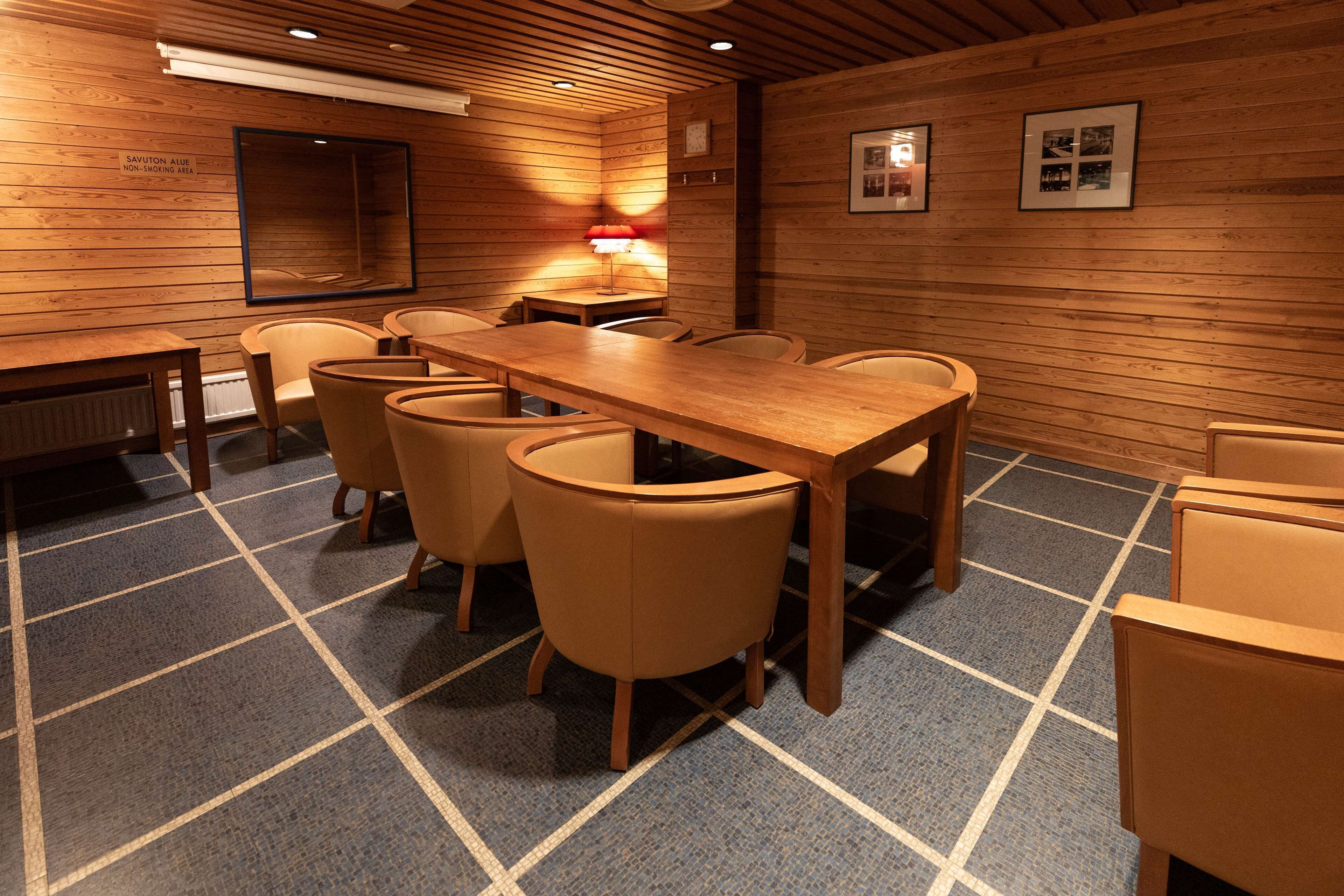 A conference room with a long table and six chairs, wooden walls, a blue carpet, and a red table lamp.