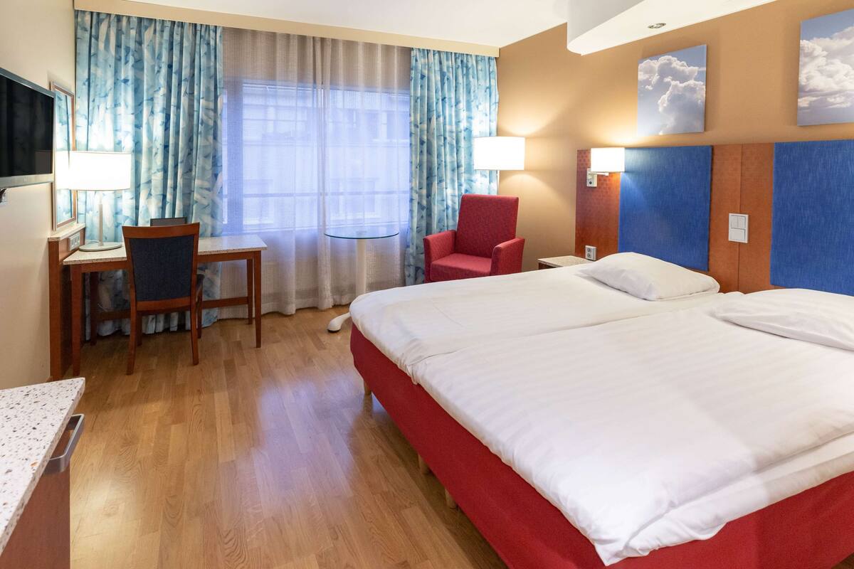 Habitación de hotel con una cama grande, un escritorio con silla, televisión y ventana con cortinas.