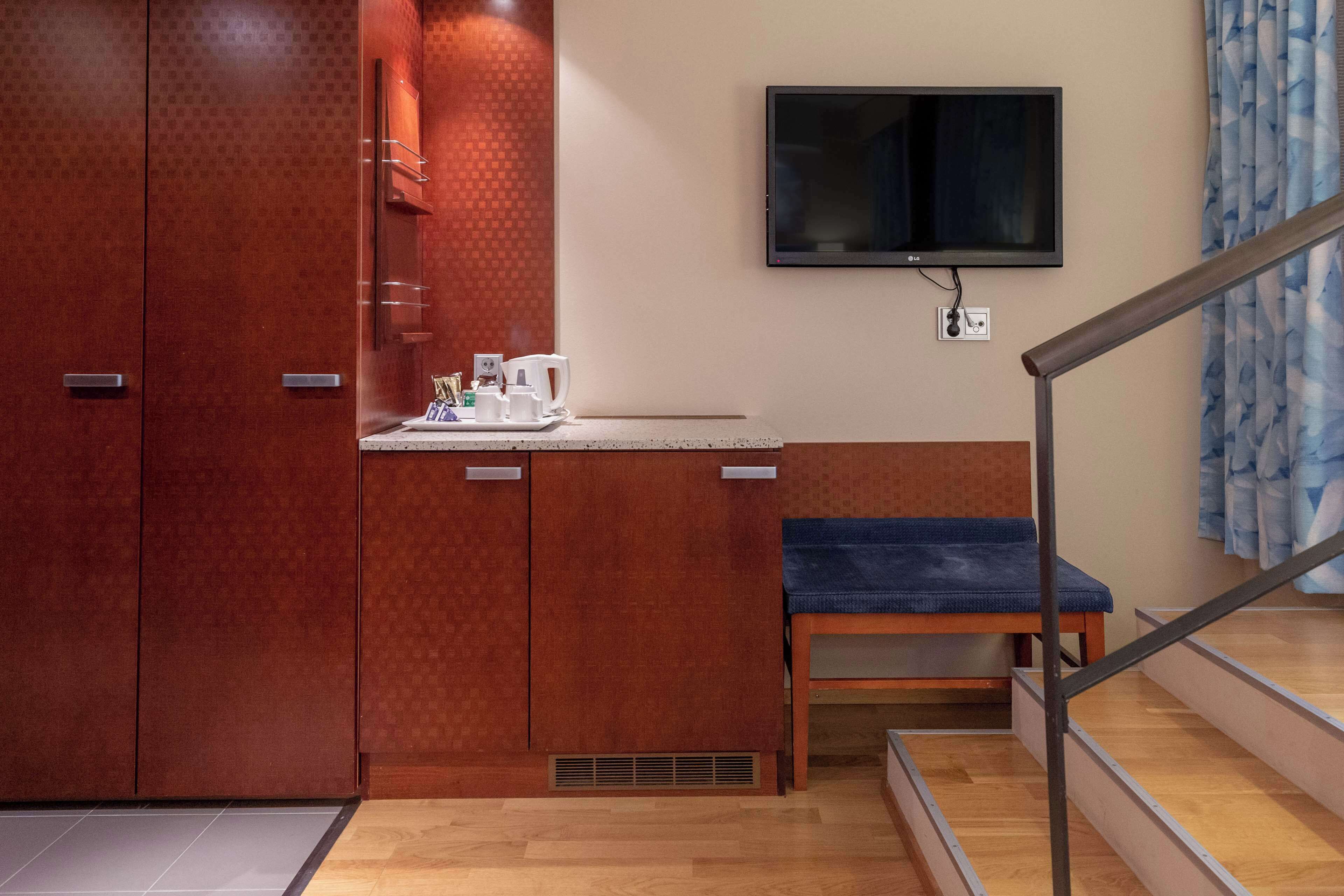 A hotel room with a wooden wardrobe, a small kitchenette with a coffee maker, a television mounted on the wall, and a wooden bench.