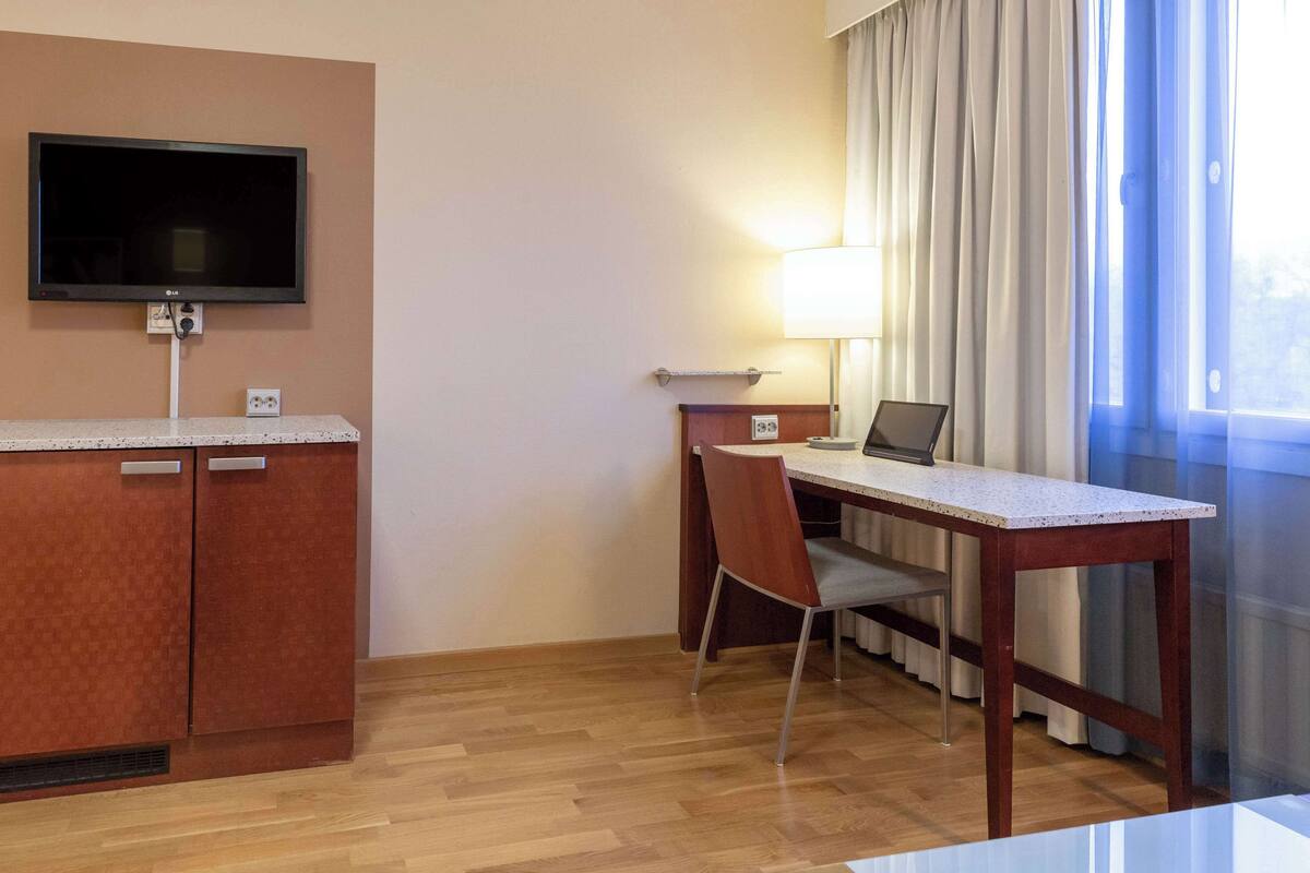 Habitación de hotel con escritorio de madera, una silla, televisor montado en la pared, lámpara y ventana con cortinas.