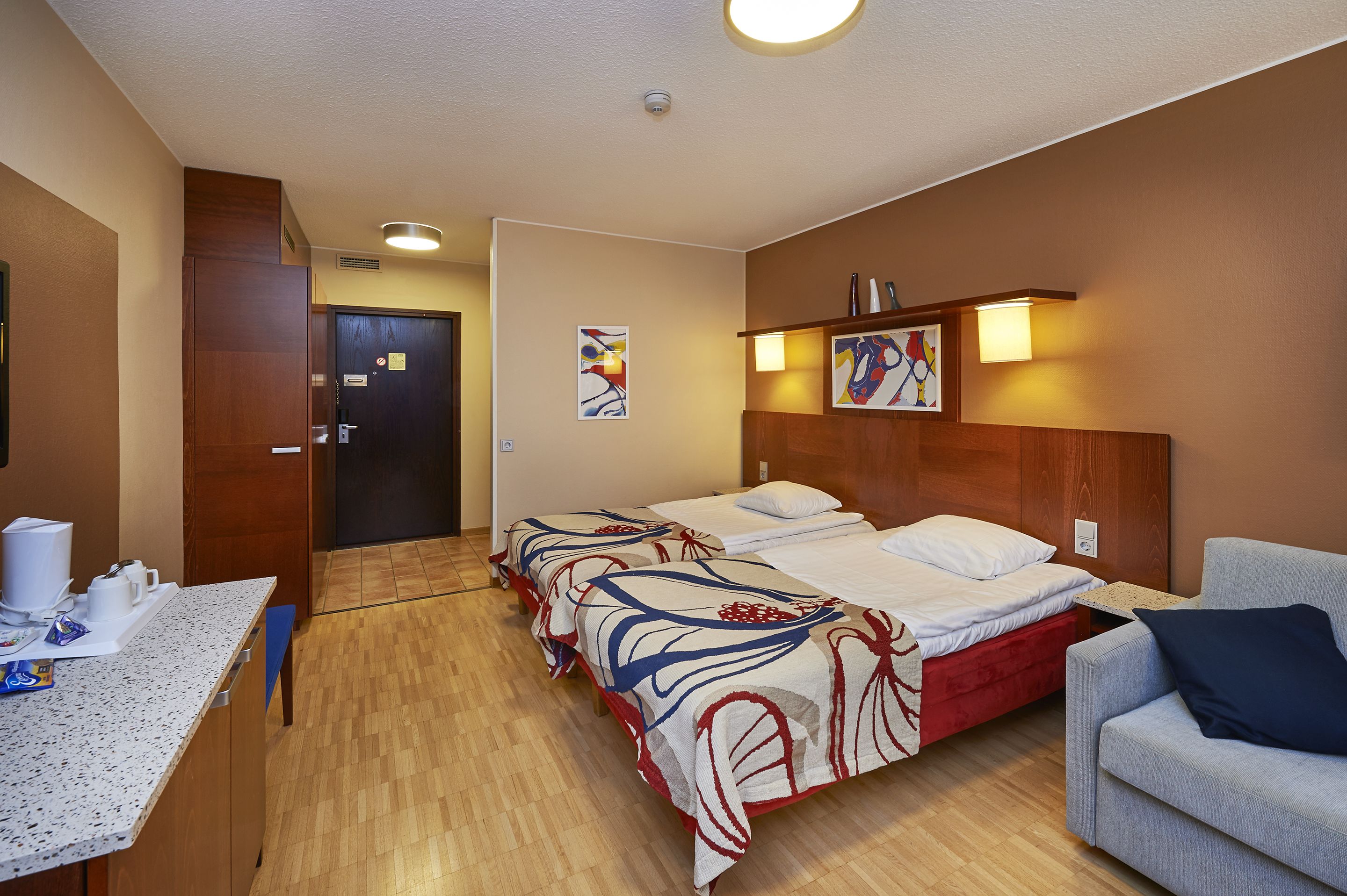 A hotel room with two beds, a desk with a chair, a sofa, and artwork on the walls.