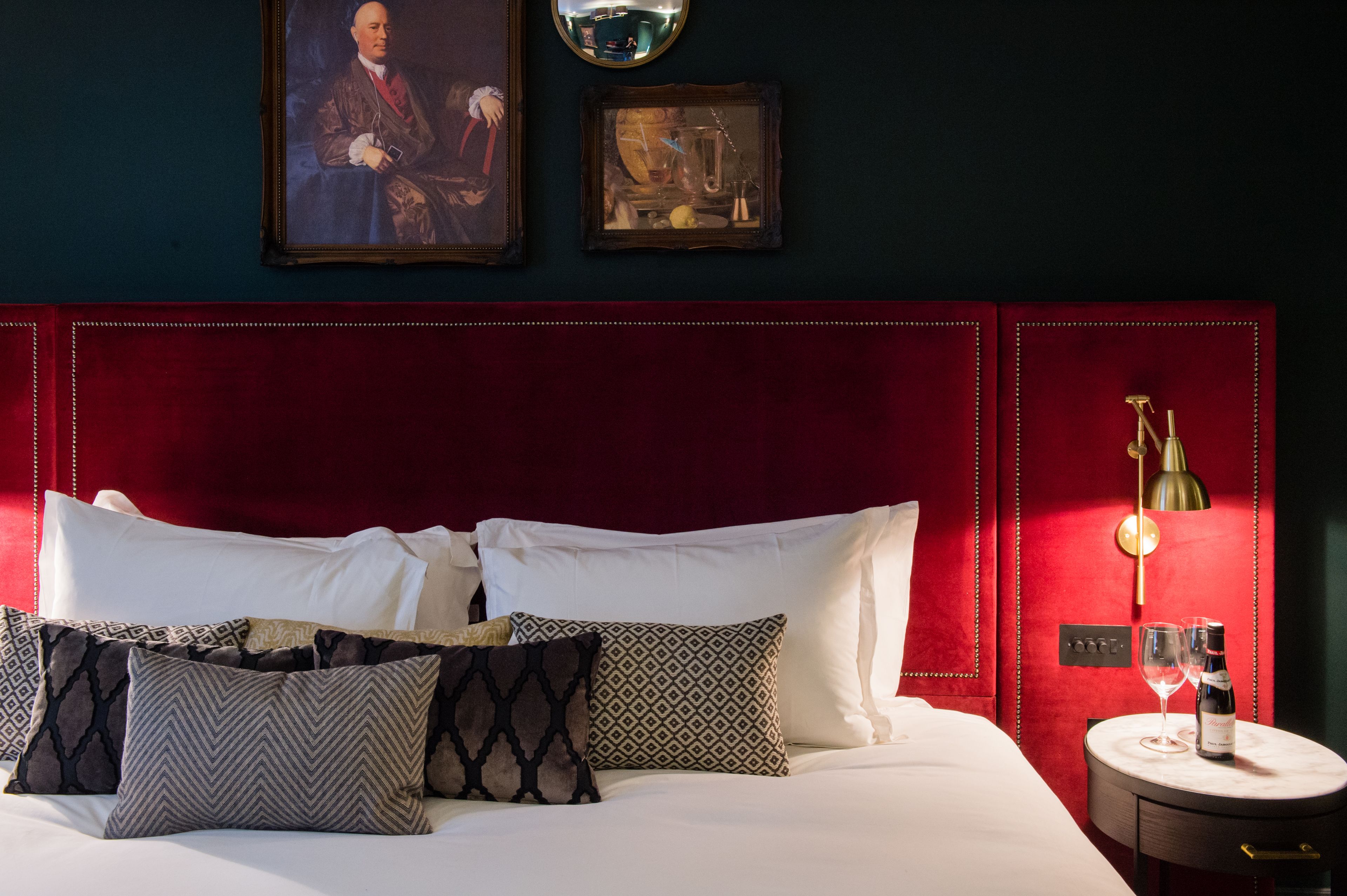 A hotel room with a red upholstered headboard, a bed with white linens, patterned pillows, a small round table with a wine bottle and glasses, and wall-mounted artwork.