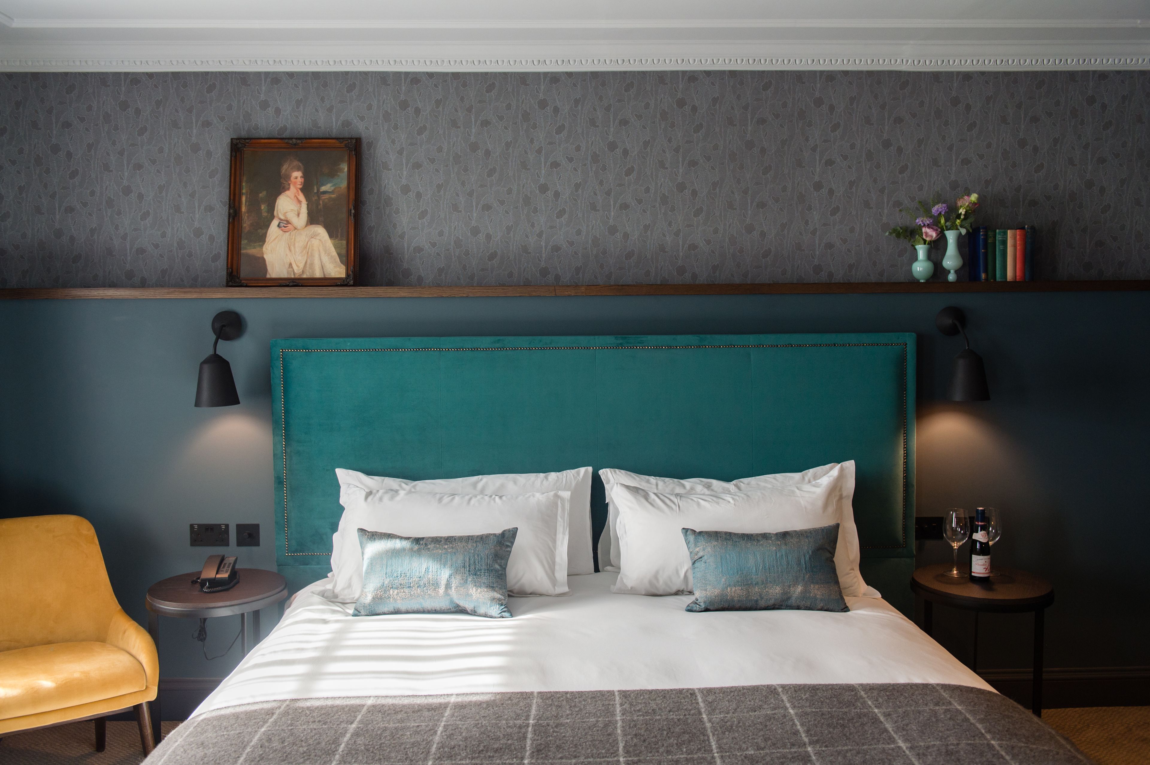 A hotel room with a teal headboard, white bedding, and a framed portrait above the bed. There are two bedside tables, one with a lamp and the other with a wine bottle and glass.