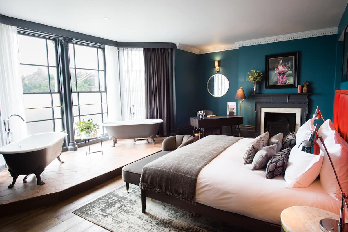 A modern bedroom with a large bed, a clawfoot bathtub, a desk, and a fireplace.