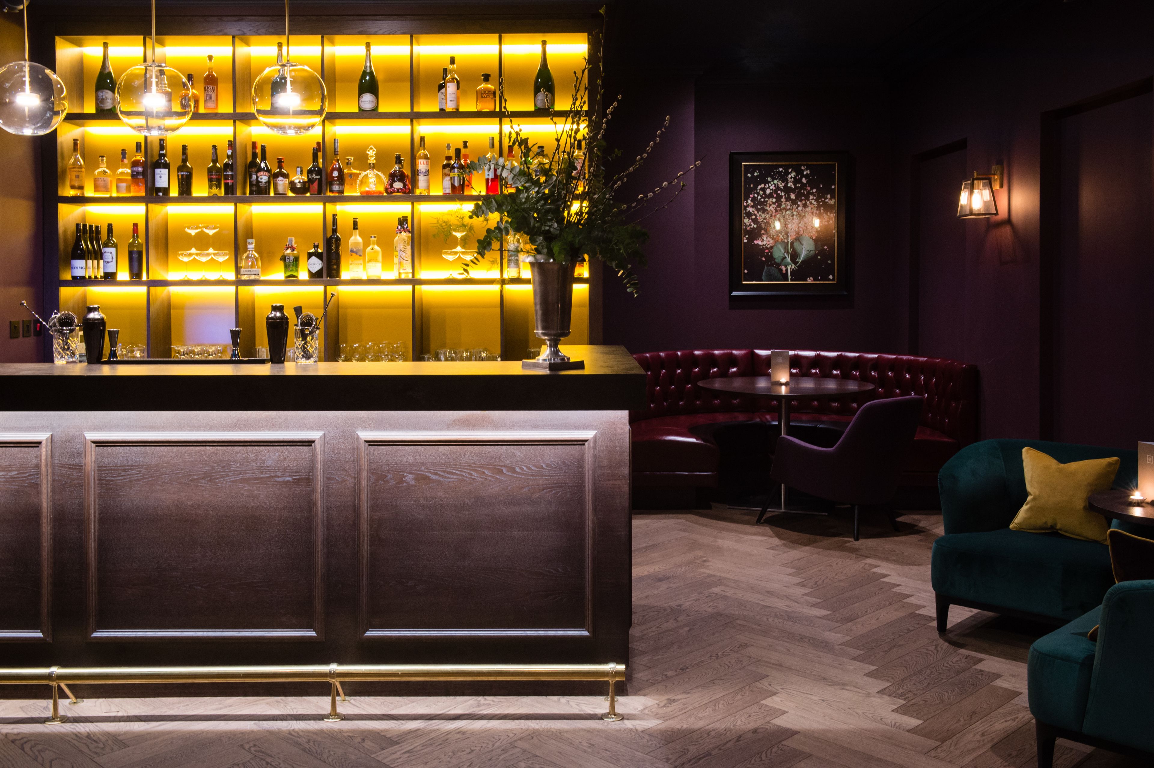 A bar area with a wooden counter, shelves of bottles, and a seating area with a red sofa and teal chairs.