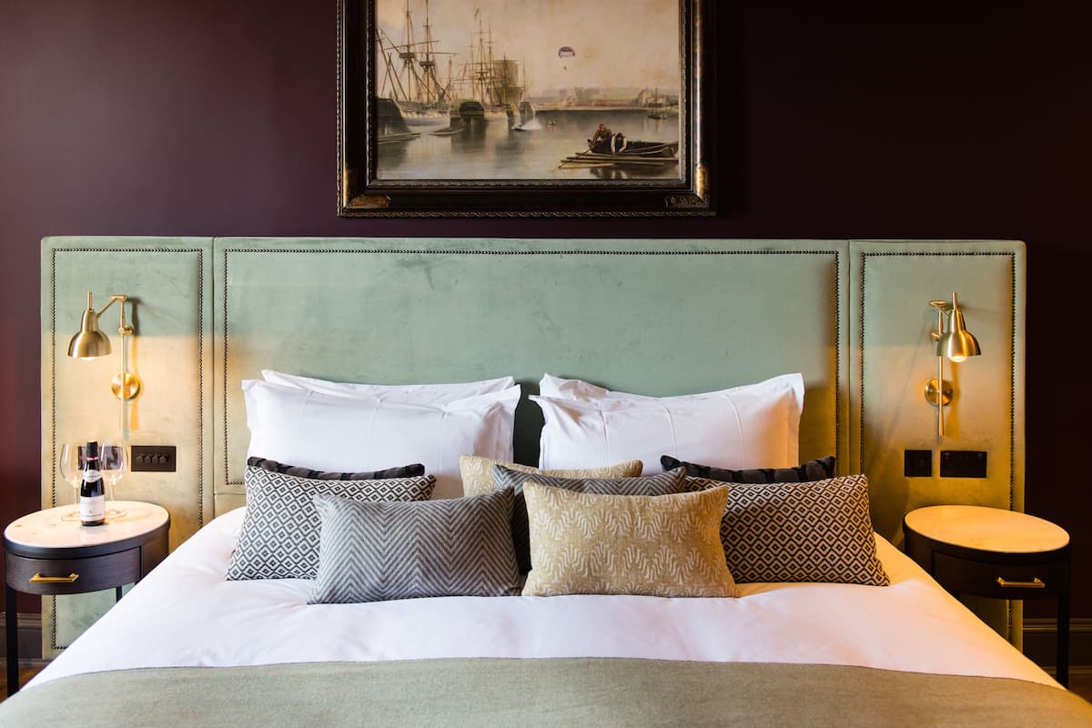 A neatly made bed with white linens and patterned pillows, flanked by two bedside tables with lamps, and a framed painting of a harbor scene above.