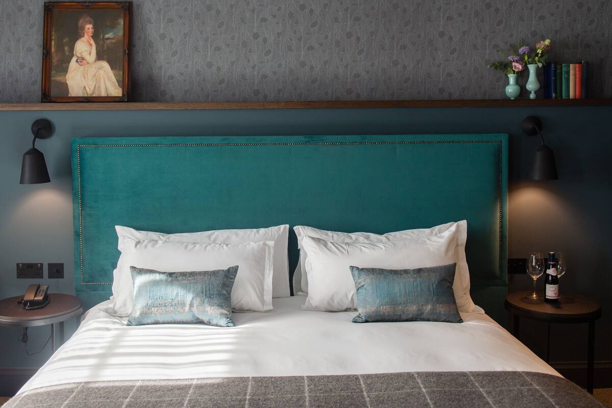 A neatly made bed with white and teal pillows, a teal headboard, and a bedside table with a wine bottle and glasses.