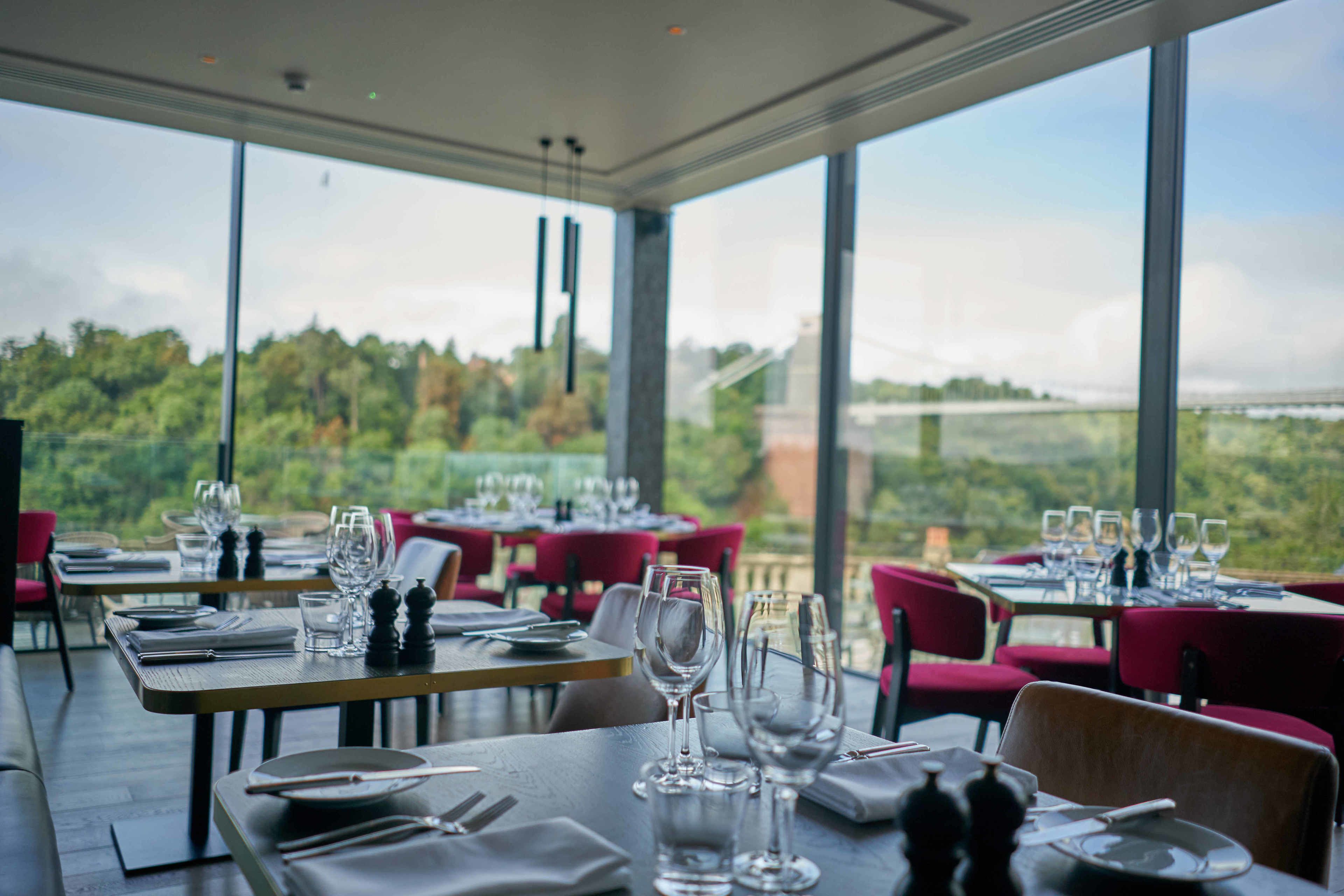 Ein Restaurant mit gedeckten Tischen, große Fenster mit Blick auf Grünflächen und eine entfernte Brücke.