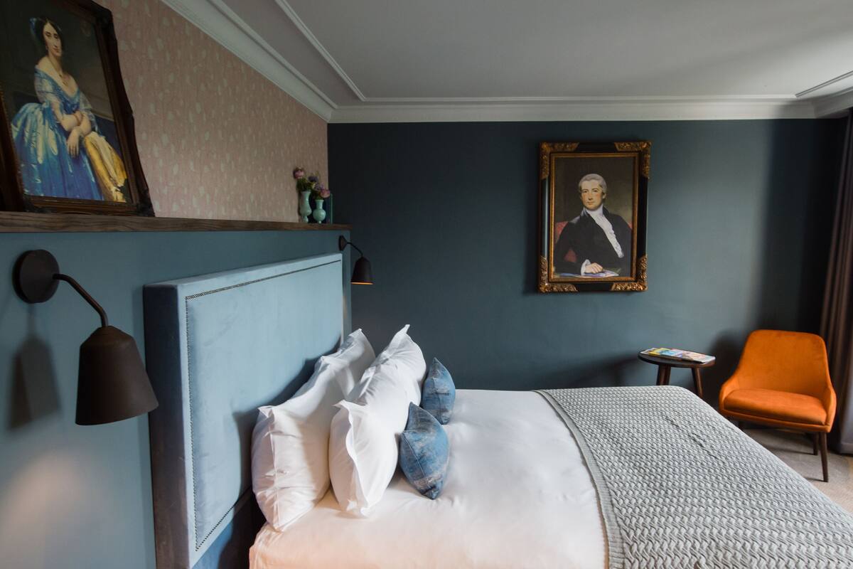A hotel room with a large bed, a nightstand, a reading lamp, a framed portrait, and an orange chair.