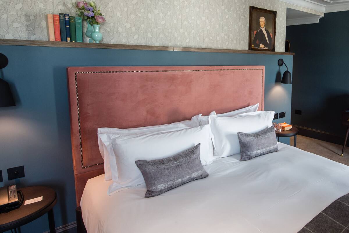 A neatly made bed with a pink headboard, white and gray pillows, and a small bedside table with a lamp.